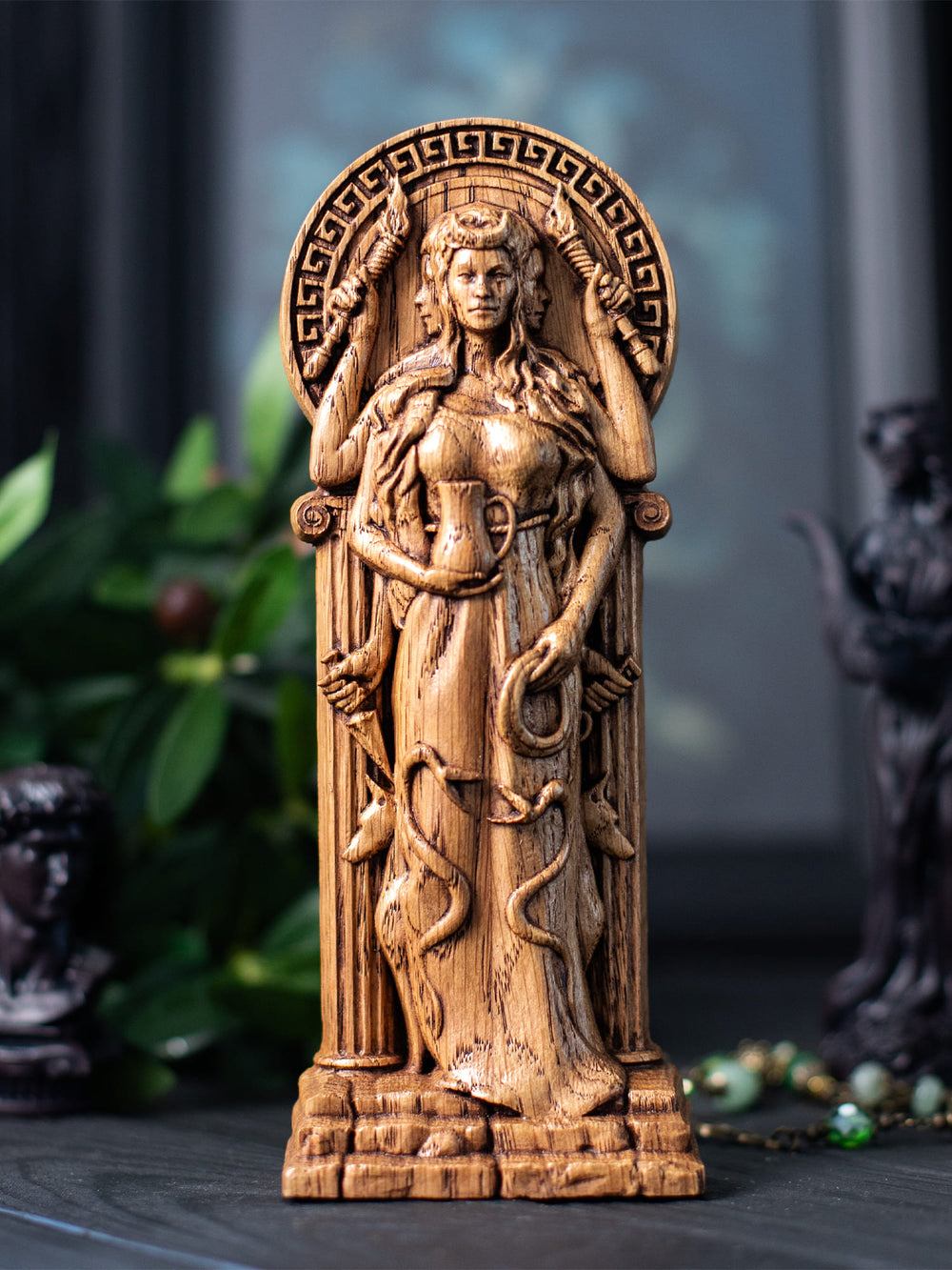 Hand-carved wooden statue of Hecate, the Greek goddess of witchcraft, crossroads, and magic. The figure is shown with multiple arms holding torches, a cup, and a key, with serpent details along the dress and small dog figures near the base. Hecate stands between classical columns beneath a circular Greek ornamented arch, creating a mystical altar-style composition in carved wood.
