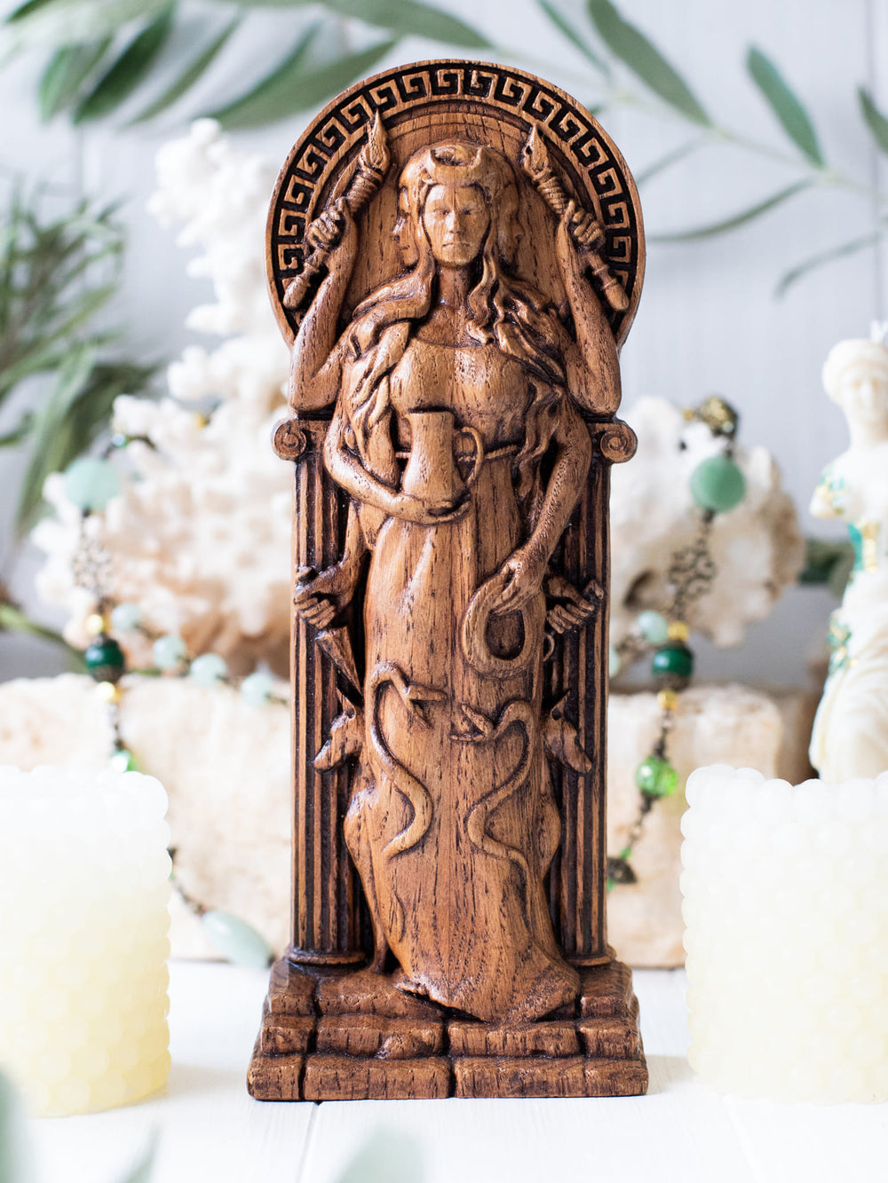 Full view of the Hekate statue from the front, highlighting its dimensions and intricate design with snakes entwined around her dress and two dogs behind her against a backdrop of ancient architecture.