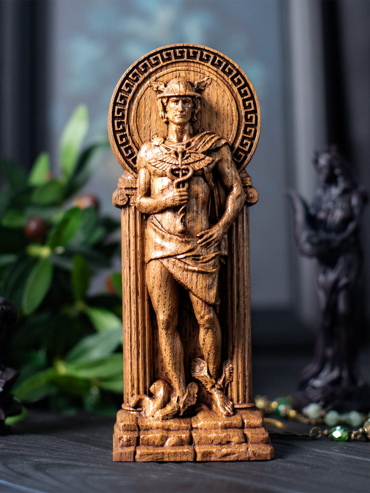 Hand-carved wooden statue of Hermes, the Greek god of travelers, trade, messages, and communication. The figure is shown wearing a winged helmet and holding a caduceus staff, standing between classical columns beneath a circular Greek ornamented arch. Small wing details near the base emphasize Hermes’ swift, divine nature. The statue is photographed on a dark surface with soft greenery and mystical decor in the background.