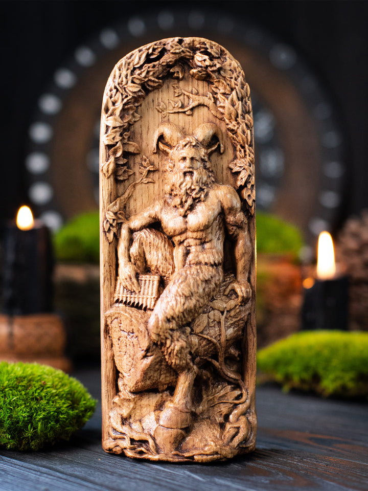 Hand-carved wooden statue of Pan, the Greek god of nature, music, and wild forests. The figure is shown with goat horns, a beard, muscular torso, and goat-like legs, holding pan flute pipes beside a leafy forest relief. The sculpture stands on a dark wooden surface with moss and candlelight in the background, creating a mystical woodland altar atmosphere.